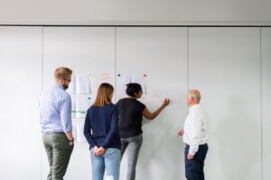 Four people standing and interacting in front of a whiteboard. One person is writing or drawing on the board while the others observe, showcasing their leadership. Papers are pinned on the whiteboard, suggesting a collaborative or brainstorming session. The room is bright and well-lit, ideal for strategy planning.