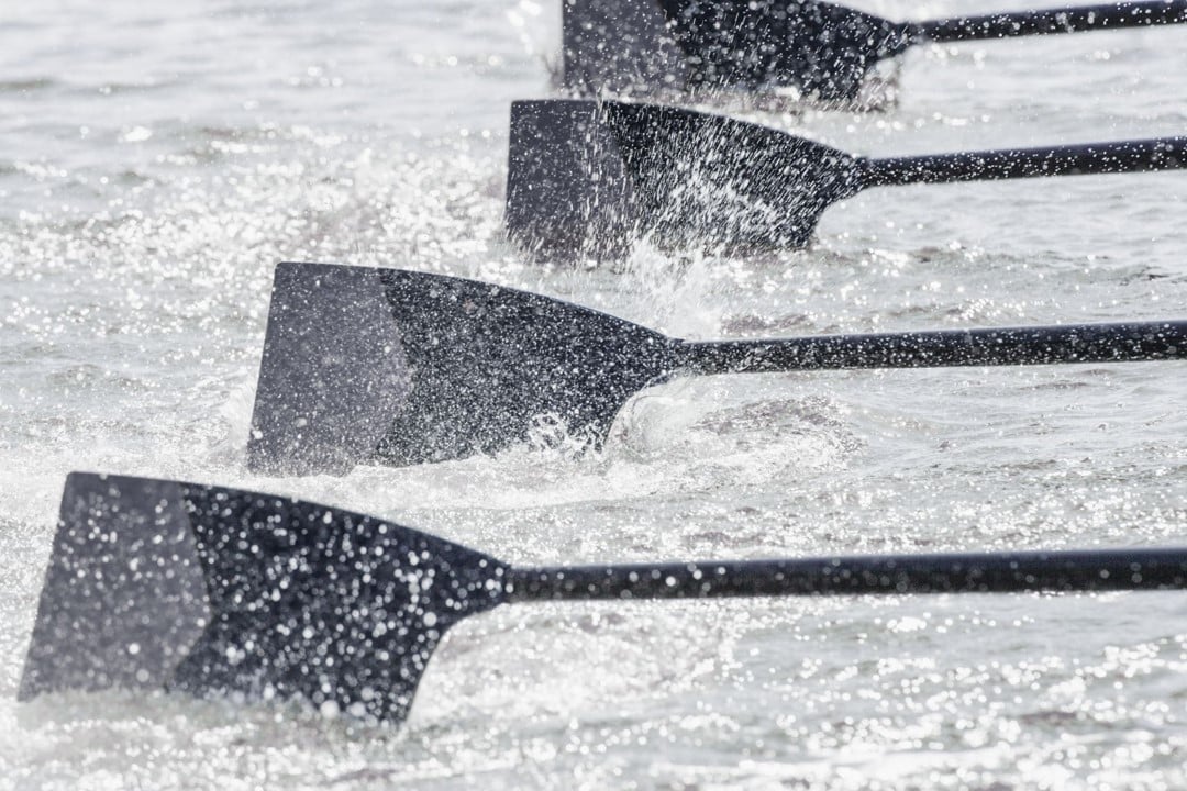 Close-up of four rowing oars in motion, splashing water as they move in unison. The paddles are black and the water appears to be choppy, indicating vigorous rowing. This scene captures the essence of teamwork and leadership, showcasing the results of effective coaching and synchronized effort.