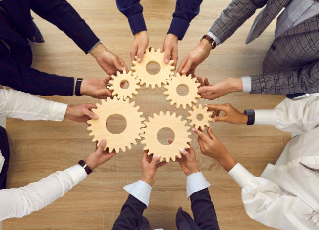 A group of people in business attire collaborate by holding interlocking wooden gears in a circle on a wooden table, symbolizing the superpower of teamwork and the vulnerability that fosters true cooperation.